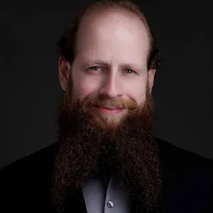 Business headshot of man with receding brown hair and full long thick brown beard wearing black suit jacket with gray dress shirt photographed against dark gray studio backdrop with dramatic professional lighting by Richard Waine Photography headshot photographer