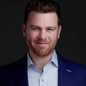 Professional headshot of Derek Lau with short styled brown hair and full reddish-brown beard wearing navy blue suit jacket over light blue dress shirt photographed against dark gray studio backdrop with dramatic professional lighting by Richard Waine Lancaster Pennsylvania headshot photographer