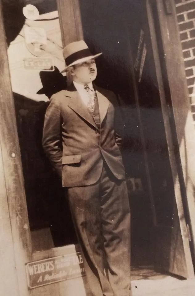 Vintage sepia-toned photograph from 1930s era showing pharmacist Abraham Krutick standing in doorway of his pharmacy wearing formal dark suit with fedora hat and mustache, brick building exterior with pharmacy signage visible, historical family photograph of immigrant pharmacist and entrepreneur during Great Depression