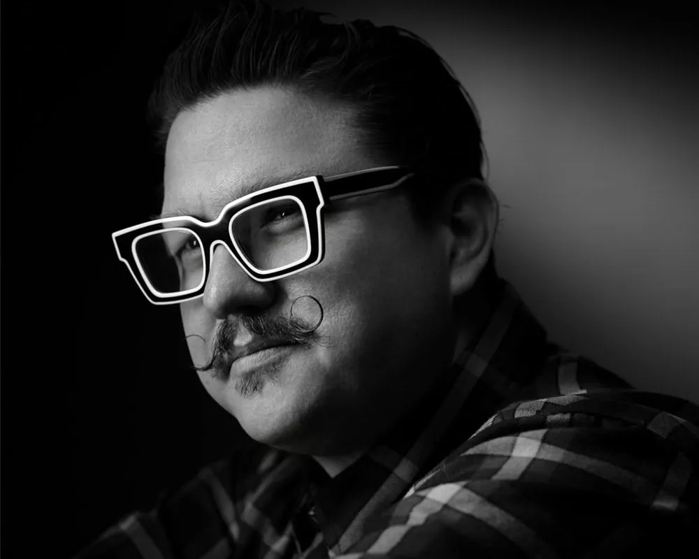 Black and white artistic portrait of Las Vegas photographer Mondo Rojas with slicked-back short dark hair wearing oversized white-framed square eyeglasses with styled handlebar mustache in checkered flannel shirt photographed with dramatic side lighting against dark gradient background