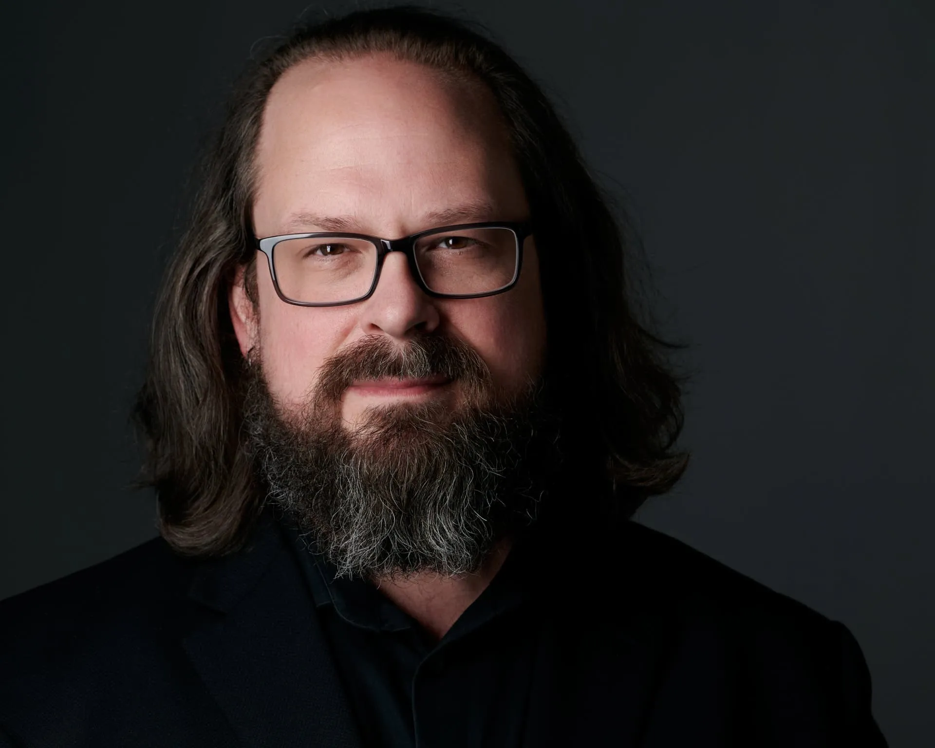 Louisville Kentucky photographer Ben Marcum professional headshot with shoulder length wavy dark brown hair, black rectangular frame glasses, full grey-streaked beard, wearing black shirt photographed on dark grey background, interview subject specializing in headshots portraits and artistic boudoir photography