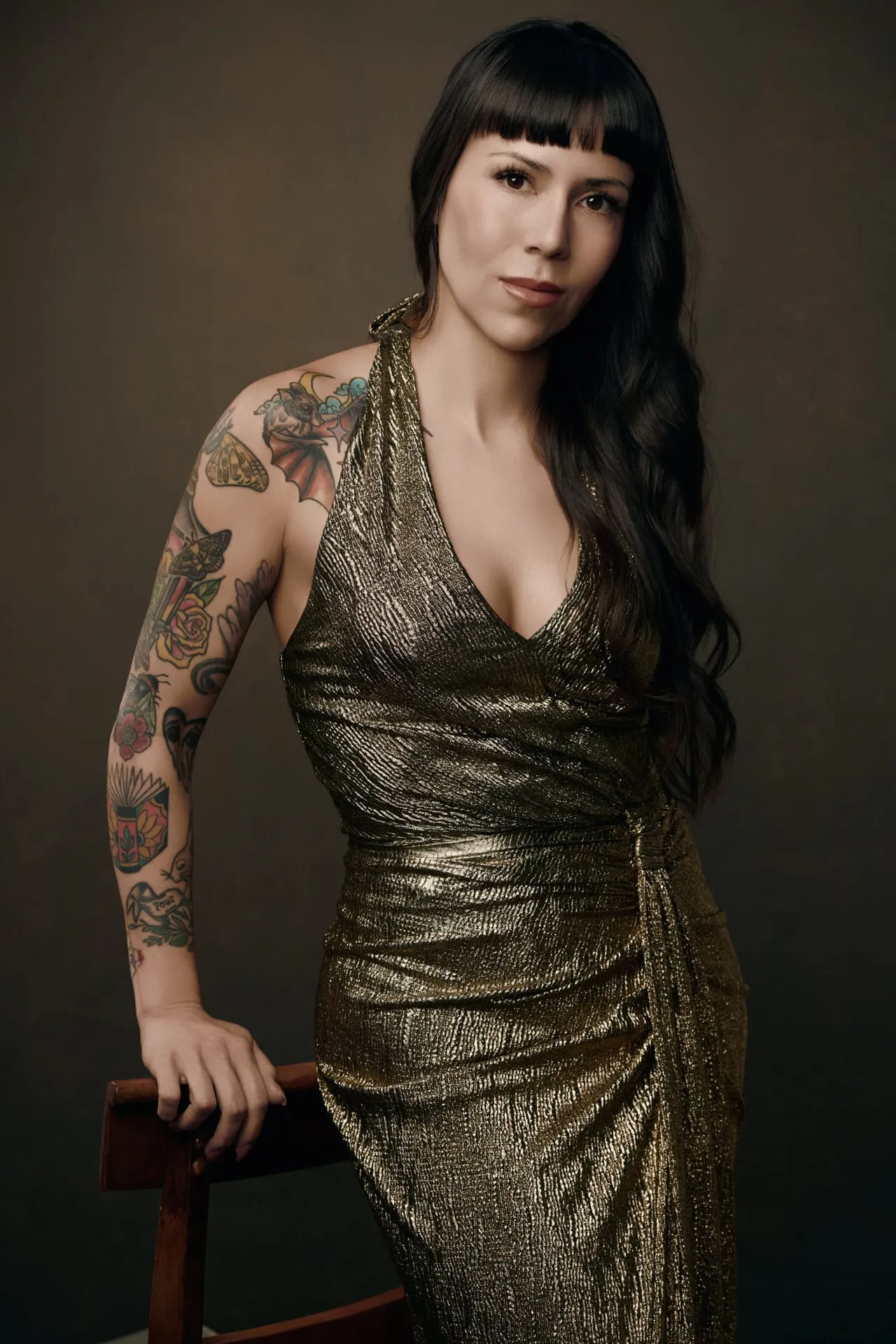 Contemporary portrait by Ben Marcum Photography featuring female with straight black hair with blunt bangs, colorful tattoo sleeve covering left arm, wearing gold metallic halter wrap dress, leaning against brown wooden chair on textured tan canvas background, Louisville Kentucky portrait photography example