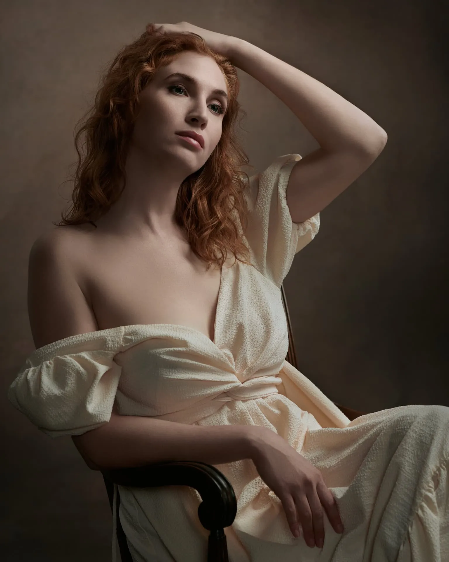 Fine art portrait by Ben Marcum Photography featuring female with long wavy red auburn hair, hand raised to head, wearing cream off-white textured robe with tie waist, seated elegant pose on dark wooden chair against warm brown textured background with Rembrandt lighting, Louisville Kentucky boudoir portrait photography example