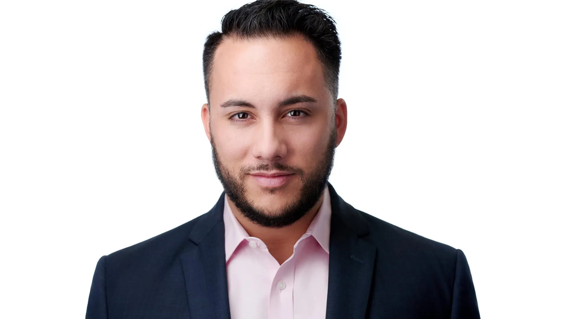 Professional corporate headshot of Italian American man with medium skin tone short dark brown hair styled with volume on top neatly trimmed dark brown beard and mustache with dark brown eyes wearing navy blue business suit jacket with notched lapels over light pink dress shirt with spread collar showing confident professional expression with subtle closed-mouth smile photographed against clean seamless white background with professional lighting demonstrating distraction-free composition by Richard Waine Photography Lancaster Pennsylvania