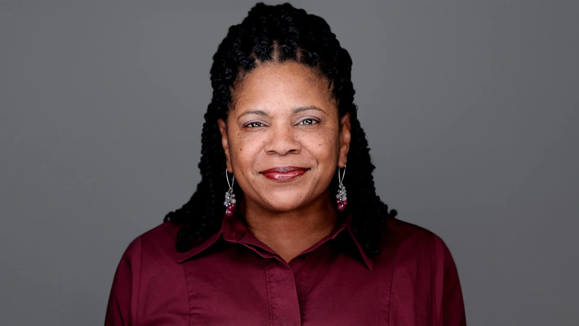 Professional headshot of African-American female with long black twisted braids pulled back, dangling silver and burgundy beaded earrings, warm smile showing teeth, wearing burgundy collared button-down shirt, photographed on grey background in Philadelphia, Pennsylvania by headshot photographer Richard Waine