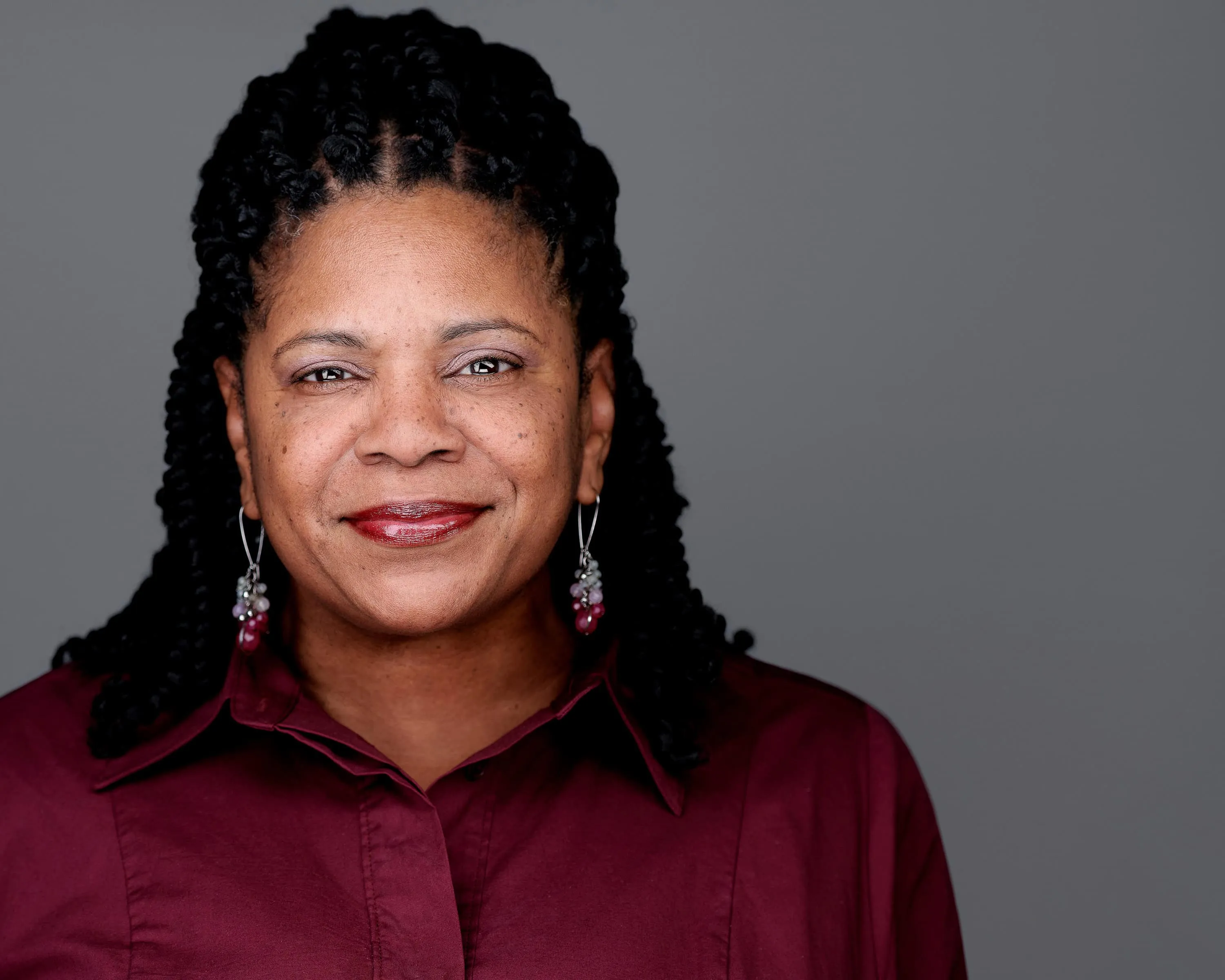 Woman with long box braids in burgundy collared shirt photographed against neutral grey studio background
