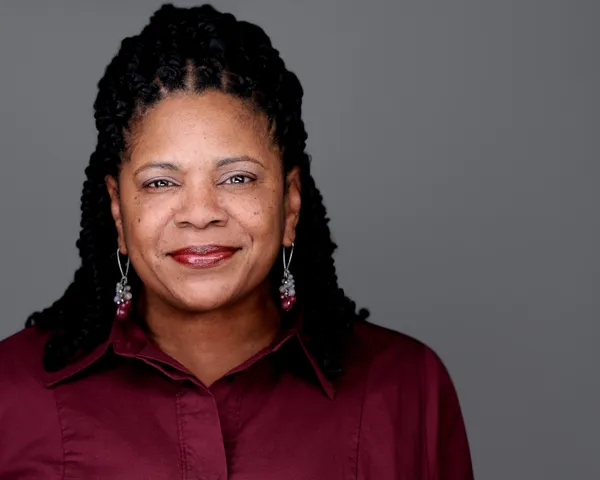 Professional LinkedIn headshot of African American woman with long braided hair in burgundy shirt, confident and poised facial expression against gray studio background, headshot made by Richard Waine in Philadelphia Pennsylvania for Chubb Insurance