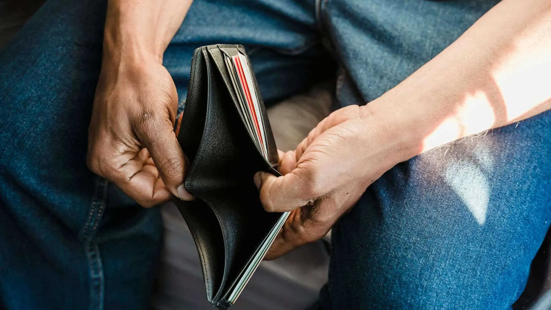 Close-up conceptual photograph of person wearing blue denim jeans holding open empty black leather bifold wallet with both hands showing no cash and only few credit cards visible inside while seated with legs visible and natural window light illuminating from right side of frame leaving left hand in shadow representing financial investment and budgeting considerations for professional headshot photography services