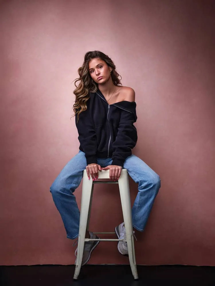Contemporary fashion portrait by Jon Erlien Photography featuring young female with long wavy highlighted blonde brown hair, wearing black zip-up hoodie jacket off one shoulder with blue denim jeans, seated on cream colored metal stool against rose pink textured gradient background, Sheboygan Wisconsin creative portrait photography example
