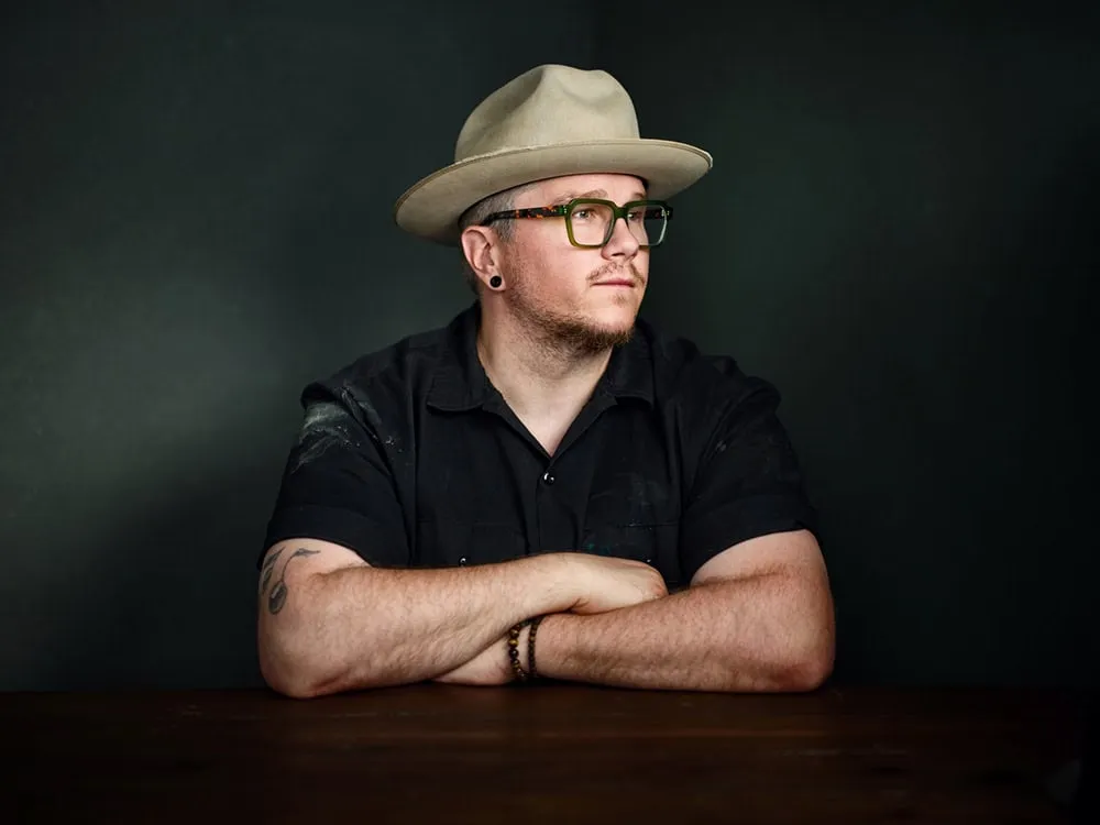 Professional portrait by Jon Erlien Photography featuring male creative with tan fedora hat, tortoise-shell frame glasses, black ear gauges, black short sleeve button-down shirt with tattoos on forearms, arms crossed leaning on dark wood table against dark teal background, Sheboygan Wisconsin contemporary portrait photography example
