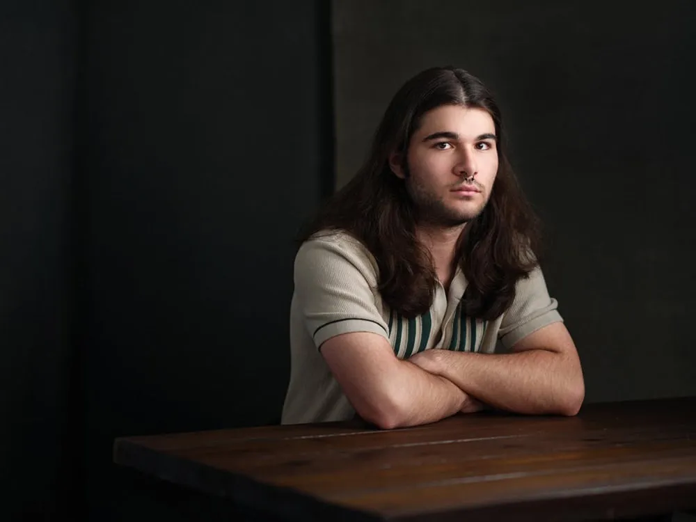 Professional portrait by Jon Erlien Photography featuring young male with long dark brown wavy hair past shoulders, trimmed beard, wearing tan beige polo shirt with teal green stripes, arms crossed leaning on dark wood table against dark background, Sheboygan Wisconsin contemporary portrait photography example