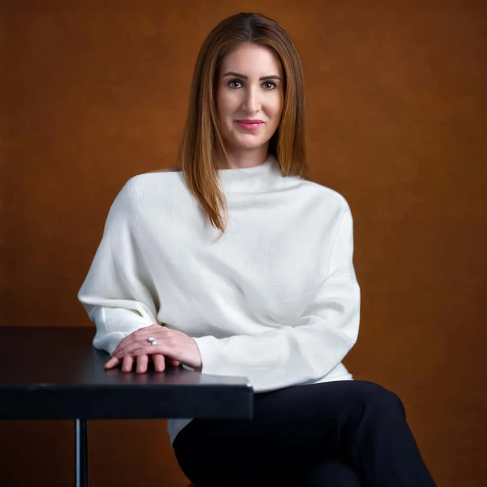 Professional business portrait by Jon Erlien Photography featuring female professional with straight shoulder-length brown hair with subtle highlights, wearing white ribbed turtleneck sweater, hands folded on black table against warm brown textured background, Sheboygan Wisconsin corporate portrait photography example
