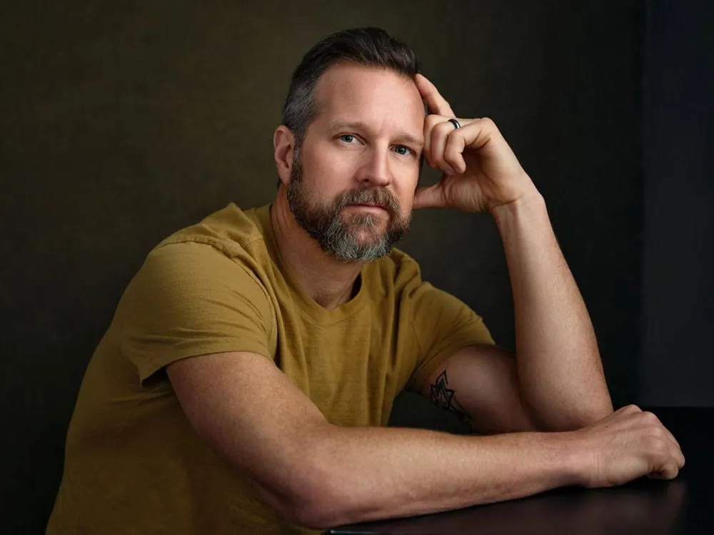 Professional portrait by Jon Erlien Photography featuring male with short dark hair styled back with grey sides, trimmed grey beard, wearing mustard yellow gold t-shirt with small tattoo on forearm, hand raised to head in thoughtful pose leaning on dark wood table against dark teal background, Sheboygan Wisconsin contemporary portrait photography example