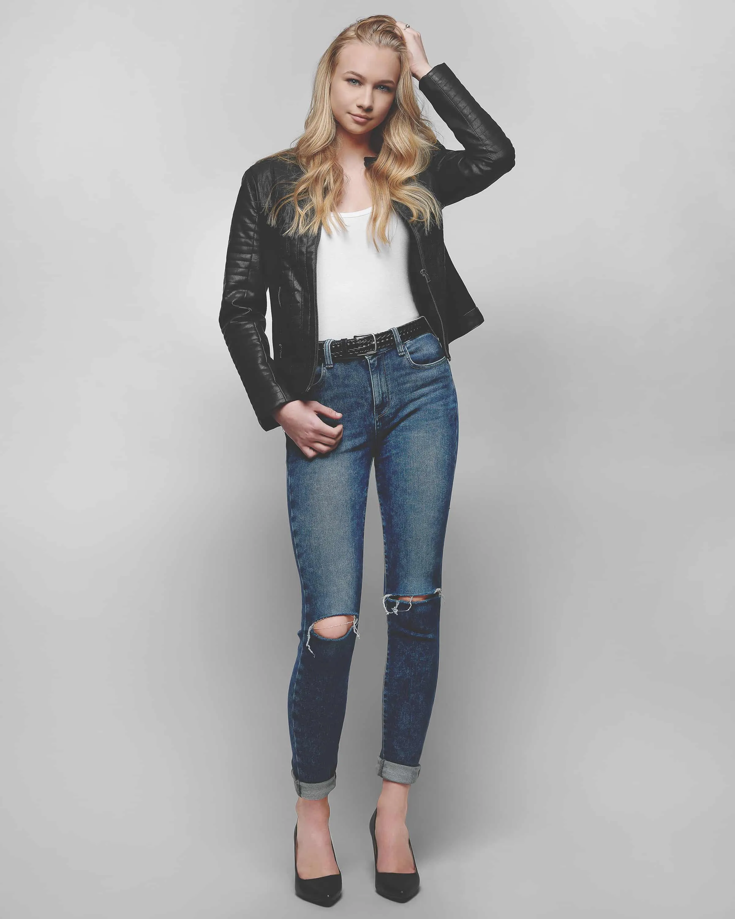 Full-length fashion modeling portrait of young female model with long blonde wavy hair wearing black leather moto jacket over white tank top with distressed rolled-cuff blue jeans brown leather belt and black pointed-toe heels photographed against neutral gray studio background by Richard Waine Photography Lancaster Pennsylvania, model portraits, modeling pictures