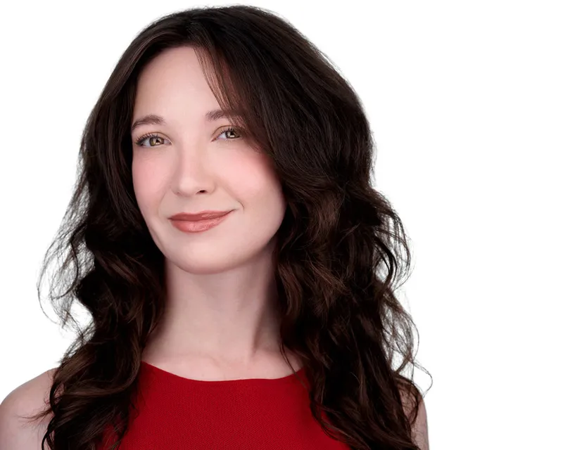 Professional actor headshot of woman with long dark brown wavy hair with caramel highlights styled with soft side-swept bangs and warm gentle smile wearing vibrant red sleeveless knit top photographed against seamless white studio backdrop with soft professional lighting by Richard Waine Photography
