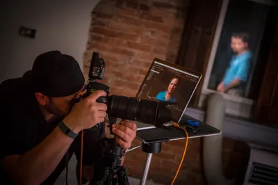 Behind-the-scenes photograph of headshot photographer Richard Waine wearing black baseball cap photographing male client in blue shirt with tethered camera setup connected to laptop against exposed brick wall at Lancaster Pennsylvania studio