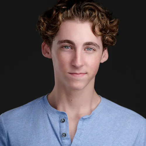 Professional actor headshot of young male with tousled medium-length brown curly hair and blue eyes with natural neutral expression wearing light blue heather henley shirt with button placket photographed against dark charcoal gray studio background by Richard Waine Photography actor headshot photographer Lancaster Pennsylvania Philadelphia Delaware