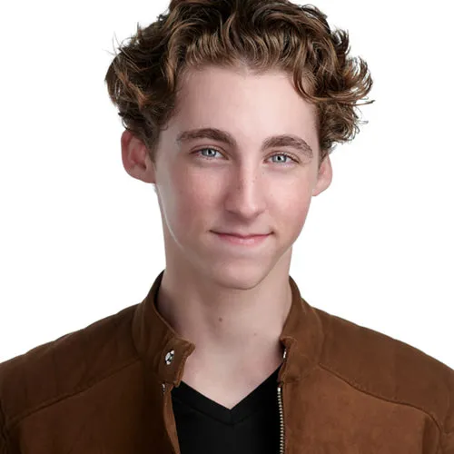 Professional actor headshot of young male with tousled medium-length brown wavy hair and blue eyes with closed subtle smile wearing tan suede bomber jacket with snap collar over black v-neck shirt photographed against white studio background by Richard Waine Photography Lancaster PA