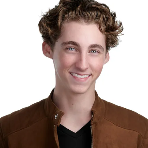 Professional actor headshot of young male with tousled medium-length brown wavy hair and bright smile wearing tan suede bomber jacket with snap collar over black v-neck shirt photographed against white studio background by Richard Waine Photography Lancaster Pennsylvania
