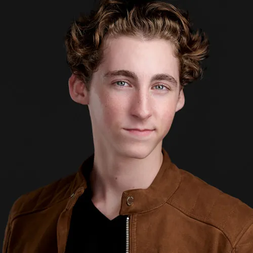 Professional actor headshot of young man with tousled medium-length brown wavy hair and blue eyes with natural light smile wearing tan suede bomber jacket with snap collar over black v-neck shirt photographed against dark charcoal gray studio backdrop with dramatic professional lighting by Richard Waine Photography actor headshot photographer Lancaster Philadelphia Pennsylvania Newark Wilmington Delaware, acting headshots lancaster, headshots for actors, Lancaster studio headshots