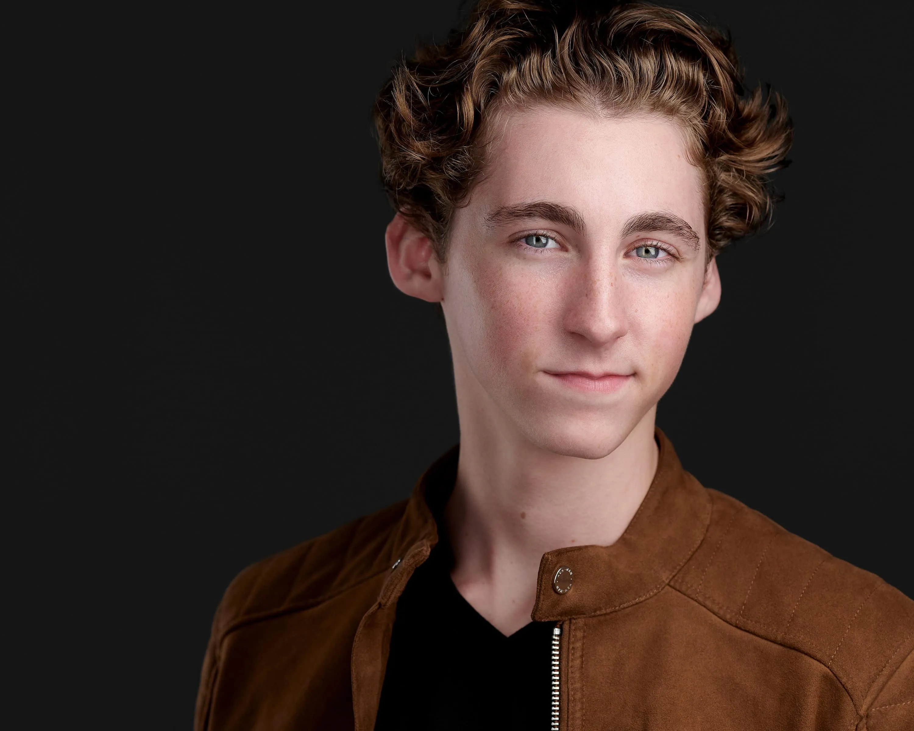 Young actor with styled wavy hair in tan suede jacket over black shirt on dramatic black studio backdrop