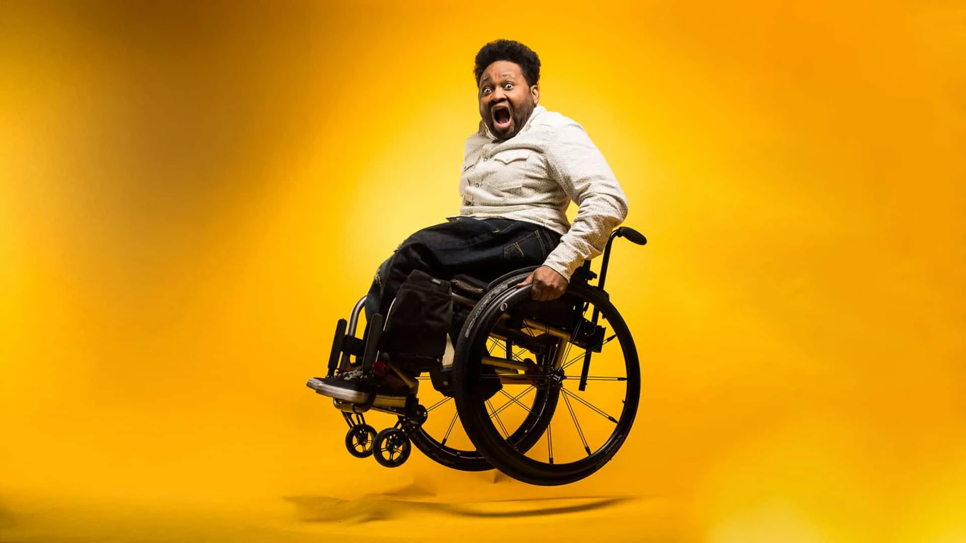 Philadelphia photographer Jaleel King creative portrait in black wheelchair with short natural hair and full beard, wearing tan beige button-down shirt with black pants, mouth open in excited joyful expression, photographed against vibrant yellow orange gradient background, disability representation in professional photography industry