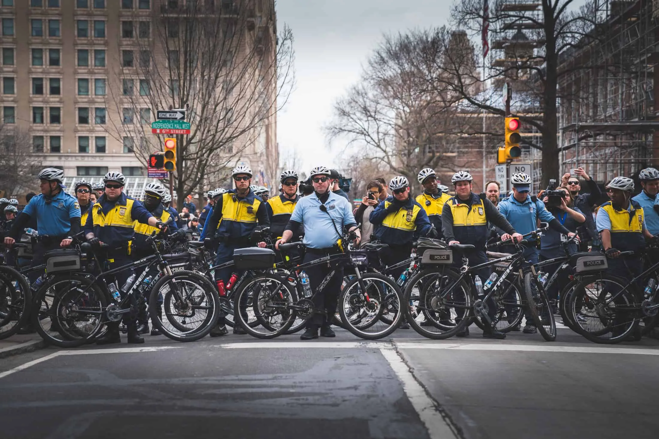 Group of police officers on bicycles lined up on urban street wearing police uniforms with badges, cycling helmets, bare winter trees and brick buildings in background, overcast sky, photograph by Jaleel King