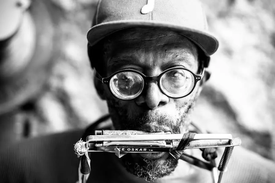 Black and white close-up portrait of male with round thick-framed eyeglasses and baseball cap with trimmed beard, holding harmonica to mouth, shallow depth of field with soft blurred background, photograph by Jaleel King