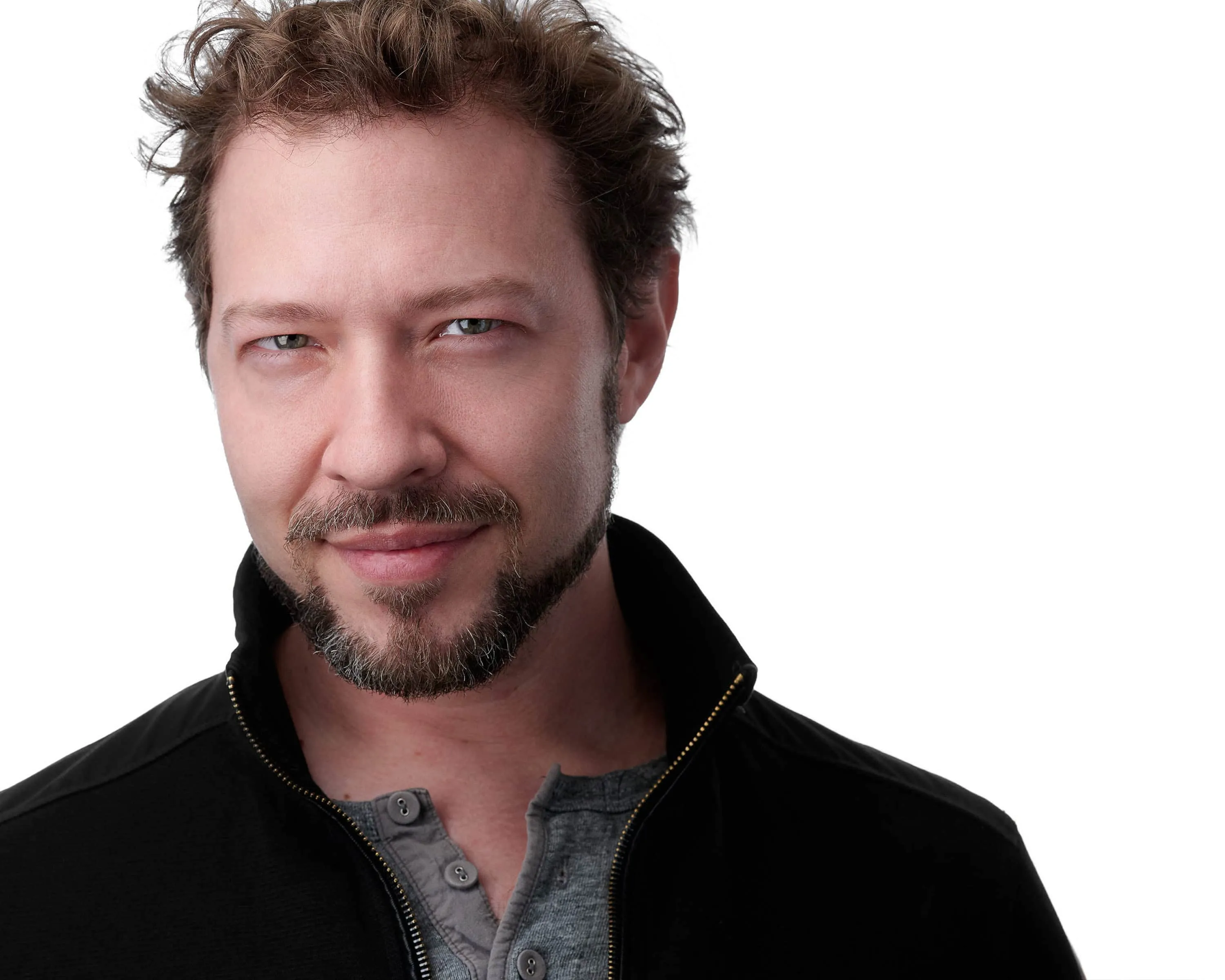 Professional actor headshot of Caucasian man with tousled medium brown hair swept back showing light colored eyes with squinch technique demonstrating confident expression well-groomed salt and pepper goatee and mustache facial hair wearing black zip-up jacket over grey henley shirt with subtle pleasant smile photographed against seamless white studio background with professional high-key lighting by Richard Waine Photography Lancaster Pennsylvania