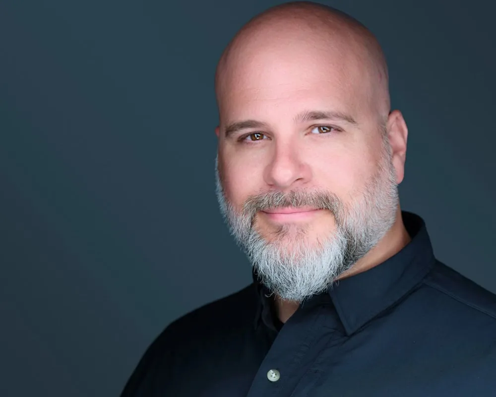 Sheboygan Wisconsin photographer Jon Erlien professional headshot with bald head, full grey beard, wearing black collared shirt photographed on dark teal grey background, interview subject specializing in headshots branding portraits and commercial photography