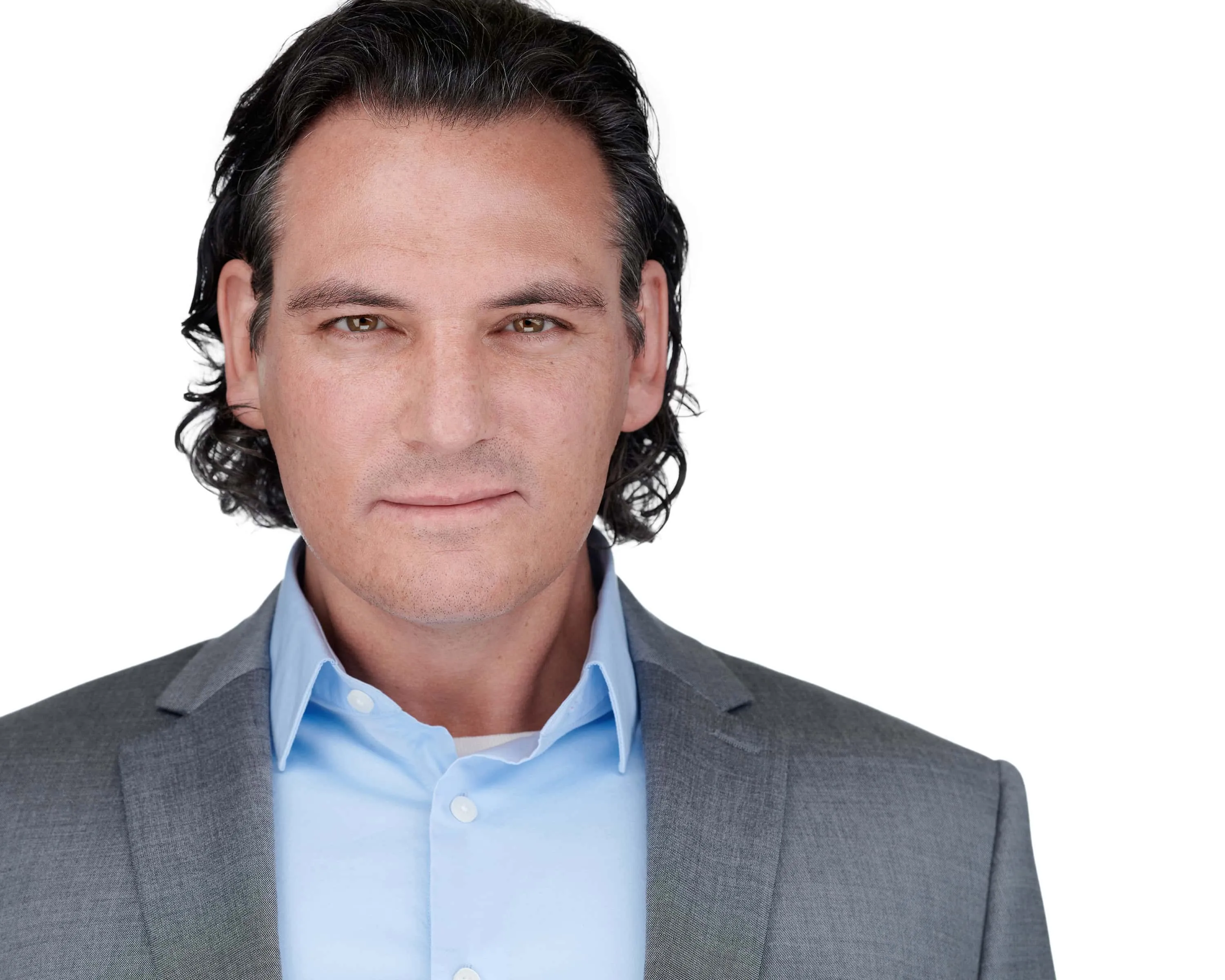 Professional headshot of male with dark brown shoulder-length wavy hair, wearing grey suit jacket over light blue collared dress shirt, photographed on seamless white background by Lancaster Pennsylvania headshot photographer Richard Waine