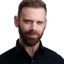 Professional business headshot of male with short dark brown hair combed back with side part and full well-groomed brown beard with defined styling wearing black polo shirt with visible collar buttons photographed against clean white studio background with professional directional lighting by Richard Waine Photography Lancaster Pennsylvania