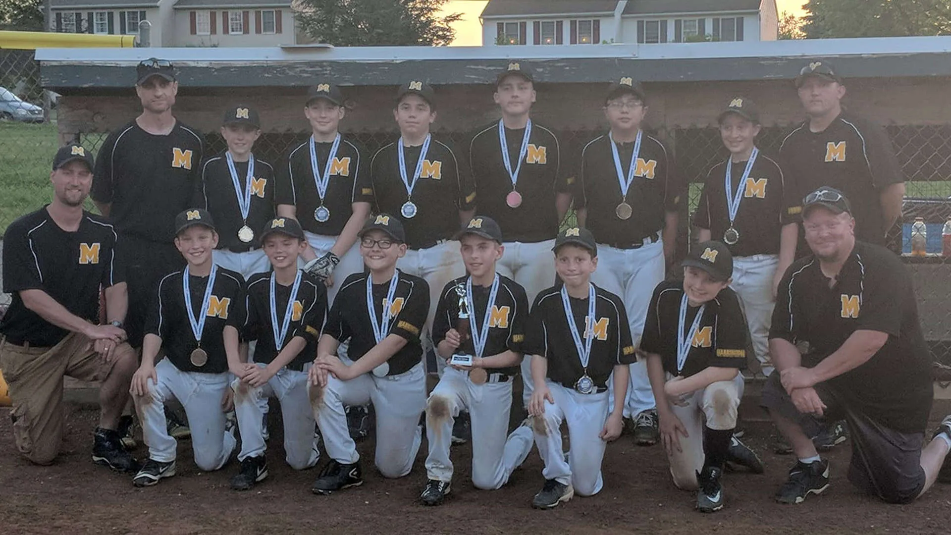 Youth baseball team photo showing Mountville Tigers players in black jerseys with gold M logo and white baseball pants, wearing medals on blue ribbons, arranged in two rows on baseball field with chain-link fence and residential buildings in background at dusk, Lancaster, Pennsylvania