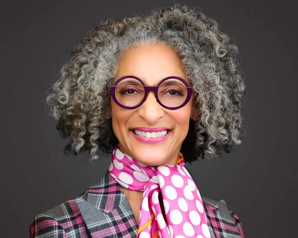 Vibrant headshot of Food Network personality Carla Hall with voluminous gray curly natural hair wearing oversized purple round frame eyeglasses bright pink polka dot silk scarf and pink gray black plaid blazer with wide smile photographed against dark charcoal studio background by Mondo Rojas