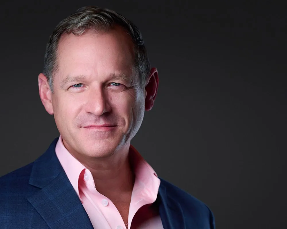Professional business headshot of male with short silver-gray hair styled wearing navy blue textured suit jacket over pink open-collar dress shirt photographed with dramatic side lighting against dark gradient background by Mondo Rojas