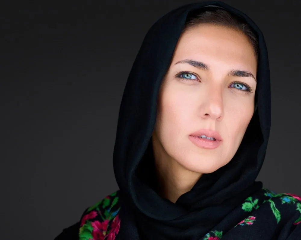 Dramatic portrait of female with blue eyes wearing black fabric hijab head covering over black top with multicolor floral embroidered detail featuring pink and green accents photographed with soft frontal lighting against dark charcoal background by Mondo Rojas