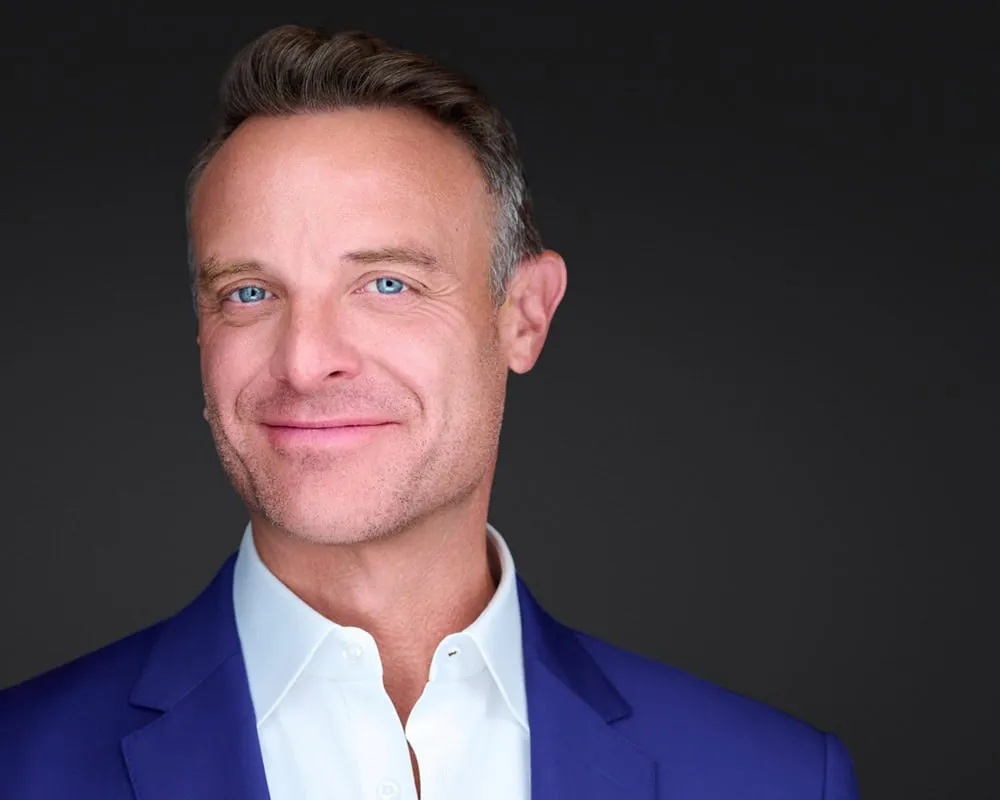Professional business headshot of male with short brown hair styled back with gray temples and blue eyes with subtle smile and light stubble wearing royal blue suit jacket over white open-collar dress shirt photographed against dark charcoal studio background by Mondo Rojas