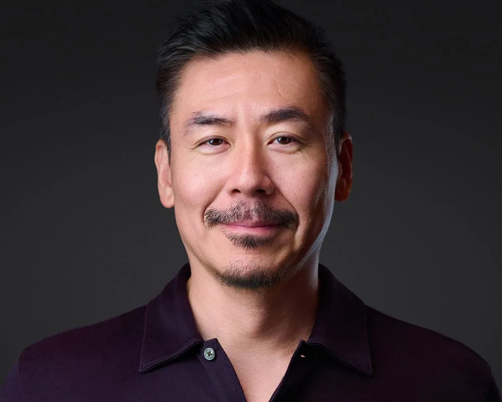 Professional headshot of Asian male with short dark hair styled back and mustache with goatee and warm smile wearing dark burgundy collared polo shirt photographed against dark charcoal studio background by Mondo Rojas