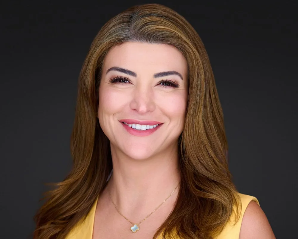 Professional headshot of female with long brown hair with side part and bright smile wearing yellow sleeveless top with gold chain necklace with turquoise clover pendant photographed against dark charcoal studio background by Mondo Rojas