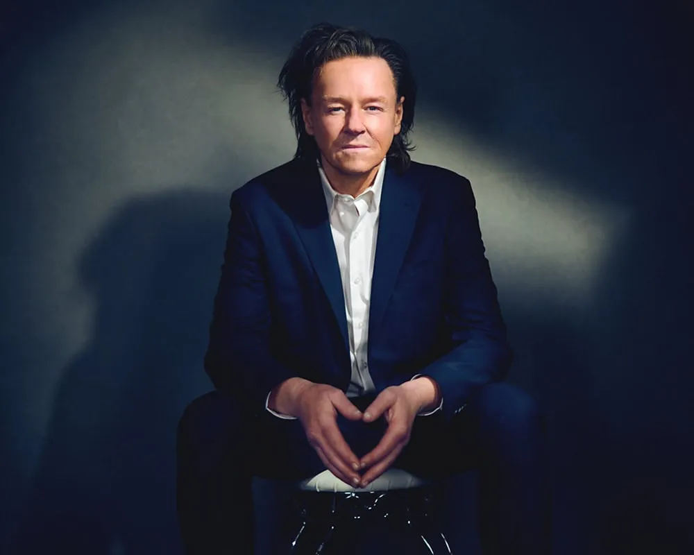 Professional portrait of male with shoulder-length dark hair styled back wearing navy blue suit jacket over white dress shirt seated with hands together photographed against dark gradient studio background with dramatic lighting by Mondo Rojas