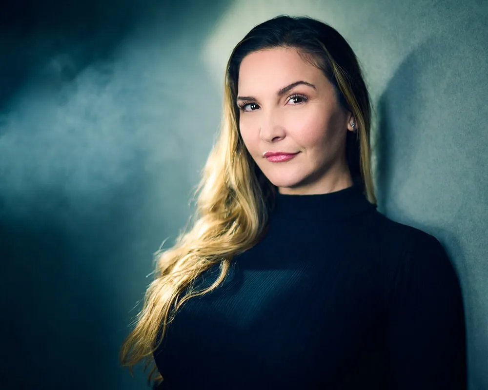 Dramatic portrait of female with long blonde highlighted hair swept to one side wearing black ribbed turtleneck sweater photographed with rim lighting against teal blue gradient studio background by Mondo Rojas