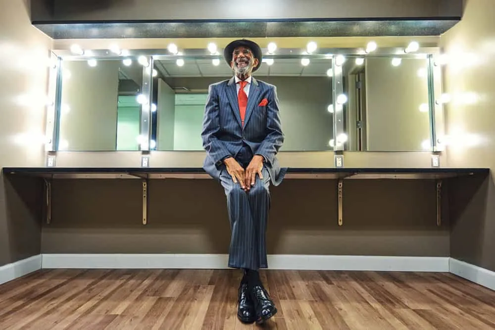 Professional portrait by Pete Coco Photography of Black male in navy blue pinstripe suit with red dress shirt, red pocket square, black fedora hat, full grey beard, sitting on dark wood counter in dressing room with large mirror surrounded by vanity lights, polished dress shoes, natural wood floor, New York portrait photography example