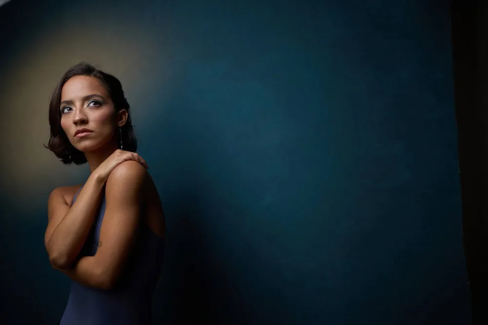 Professional portrait by Pete Coco Photography featuring female with shoulder-length dark brown hair, bare shoulders in navy sleeveless top, hand touching shoulder, photographed against dark teal blue textured background with dramatic Rembrandt lighting, New York portrait photography example