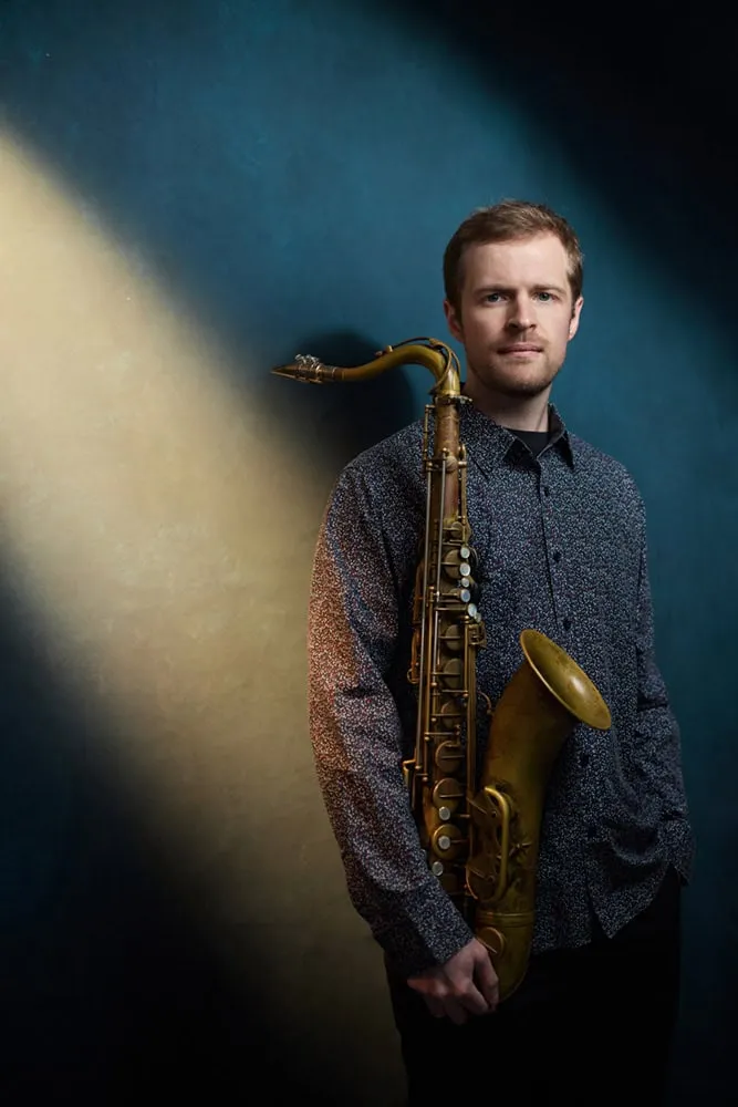 Professional musician portrait by Pete Coco Photography featuring male jazz saxophonist with short dark brown hair, trimmed beard, wearing navy blue speckled long-sleeve button-down shirt, holding brass tenor saxophone at shoulder, photographed against dramatic dark teal blue background with warm golden light streak, New York music photography example