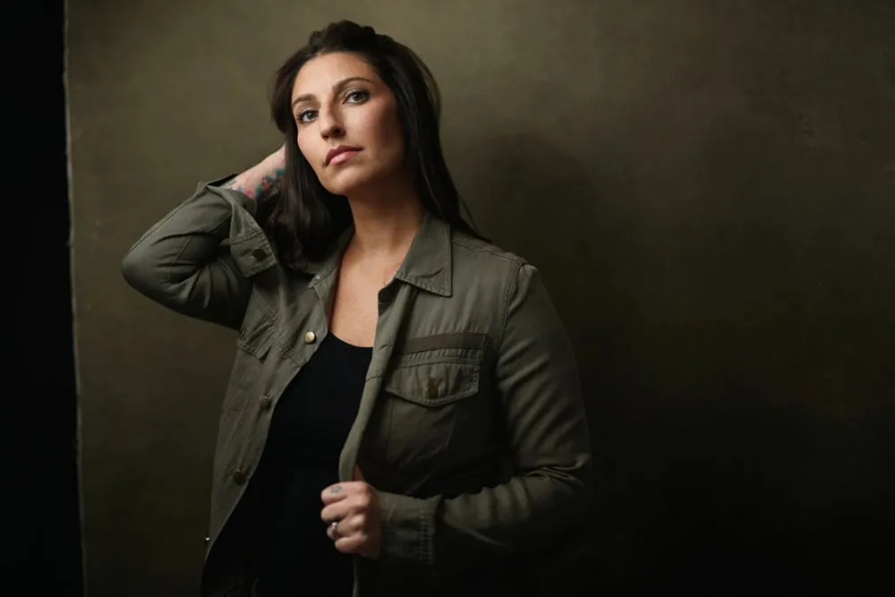 Professional portrait by Pete Coco Photography featuring female with long straight dark brown hair, visible tattoo on left forearm, wearing olive green military-style jacket over black v-neck top, hand raised to hair, photographed against dark olive green textured background with dramatic lighting, New York portrait photography example