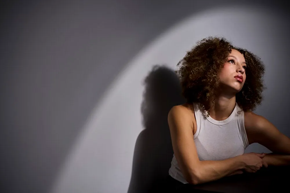 Professional portrait by Pete Coco Photography featuring Black female with voluminous curly afro hair, wearing white ribbed tank top, looking up with hands clasped, photographed against grey background with dramatic diagonal light beam creating dynamic shadow, New York portrait photography example
