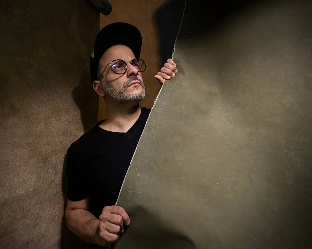 New York photographer Pete Coco creative self-portrait with oversized round tortoise-shell eyeglasses, grey beard stubble, all black outfit peeking around textured tan beige wall with dramatic shadow lighting, interview subject specializing in headshots portraits and music photography
