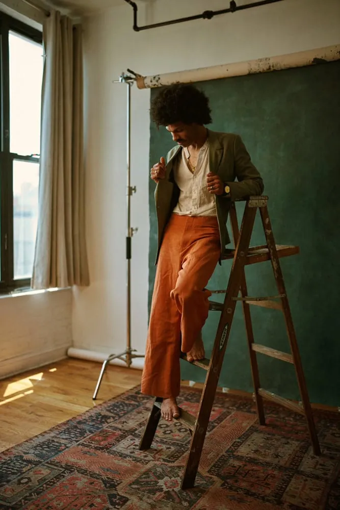 Creative lifestyle portrait by Pete Coco Photography of Black male with voluminous natural afro hairstyle, wearing olive green blazer over ivory white v-neck shirt with burnt orange rust colored pants, standing on vintage Persian rug next to wooden ladder against teal green painted wall in natural light-filled studio with windows, New York environmental portrait photography example