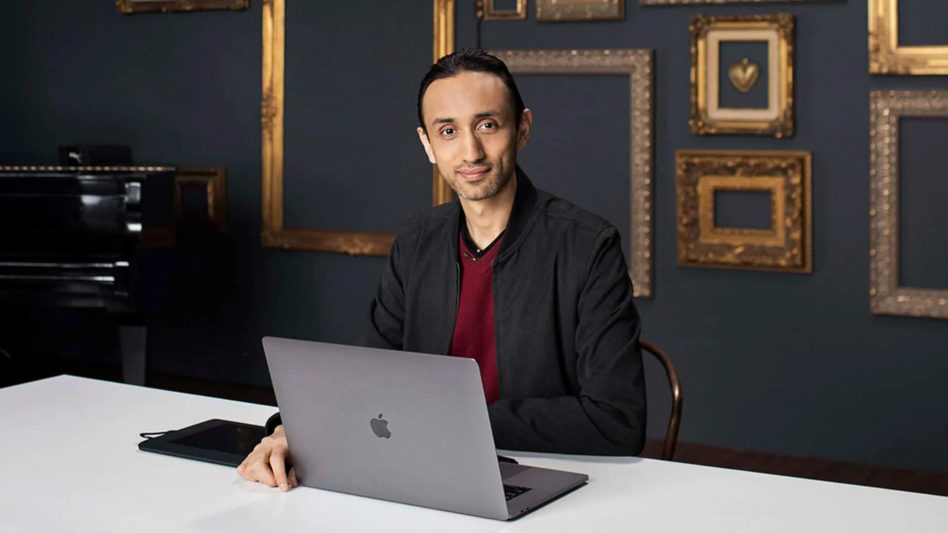 Professional retoucher Pratik Naik sitting at desk with MacBook laptop wearing dark grey blazer over burgundy shirt, slicked back dark hair, trimmed beard, confident professional expression, photographed in workspace with ornate gold picture frames on dark blue wall background, commercial photography behind the scenes