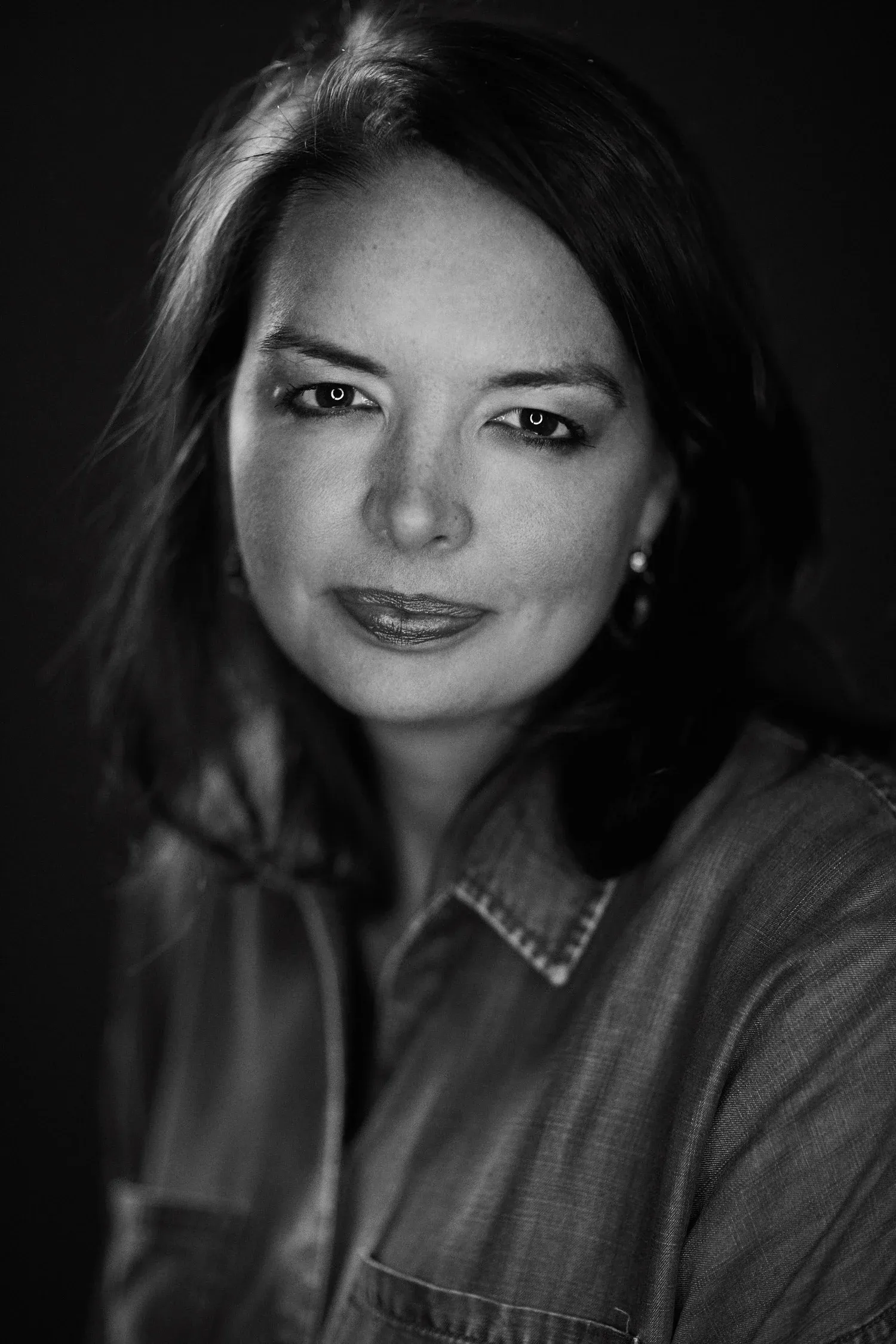 Classic black and white portrait of female professional with shoulder-length wavy hair, pearl earring, subtle smile, wearing collared denim chambray shirt, timeless sophisticated headshot style, professional skin retouching by Pratik Naik for photographer Jeremy Cowart